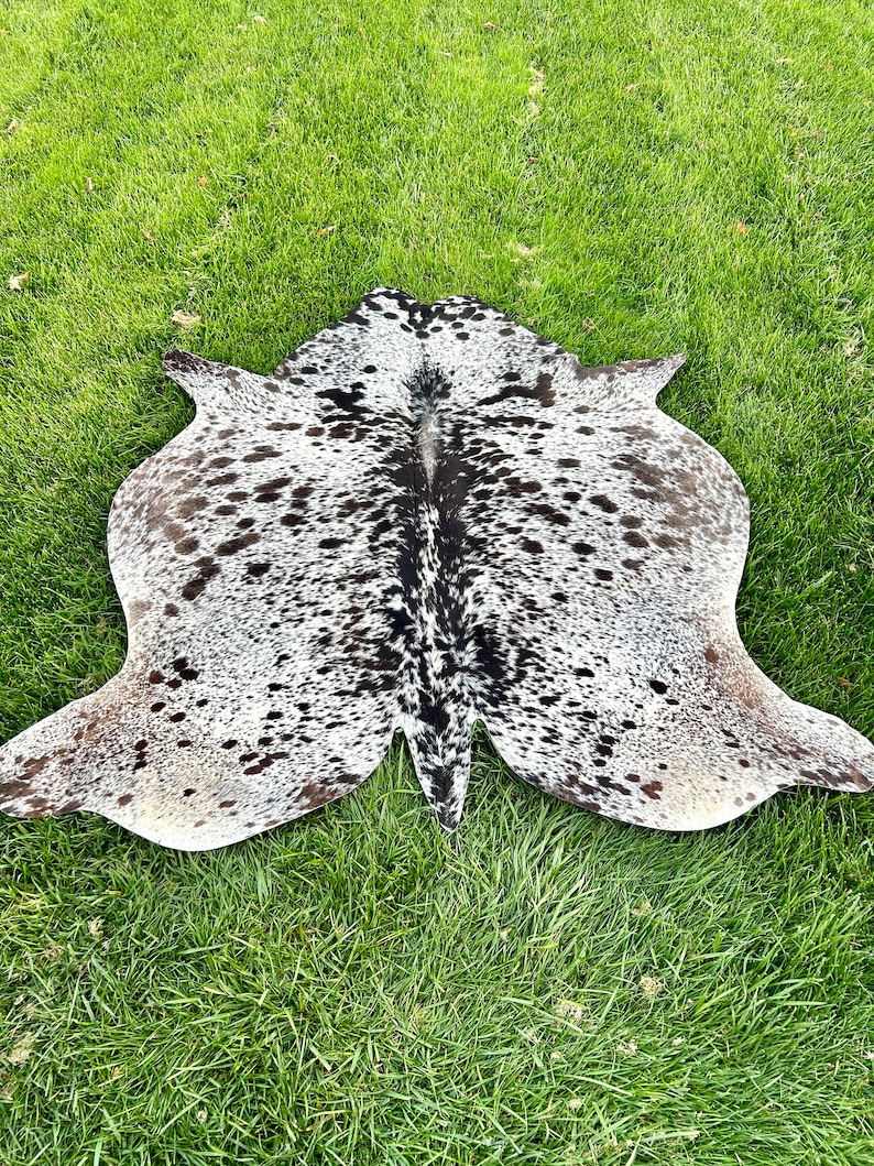 以下が含まれることがあります： 白地をベースに黒と茶色の斑点模様が施されたカウハイドラグ。 ラグは不規則な形状で、縁が伸びており、緑の草の上に置かれています。 革は柔らかく、質感のある外観をしています。