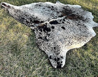 以下が含まれることがあります： 自然で不規則な形のカウハイドラグ。ラグは黒と白の斑点とパッチのパターンを特徴とし、印象的なコントラストを生み出しています。ラグは緑の草の上に置かれています。