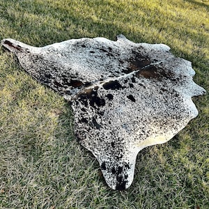 以下が含まれることがあります： 自然な不規則な形状の牛革ラグ。ラグは、黒と白の斑点とパッチのパターンで、濃い茶色とタンの色合いが混ざっています。ラグは緑の草の上に置かれています。