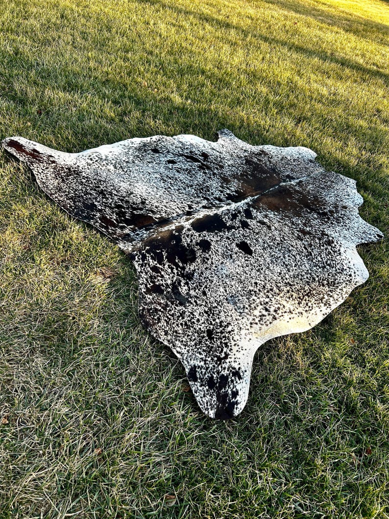 以下が含まれることがあります： 自然な不規則な形状の牛革ラグ。ラグは、黒と白の斑点とパッチのパターンで、濃い茶色とタンの色合いが混ざっています。ラグは緑の草の上に置かれています。