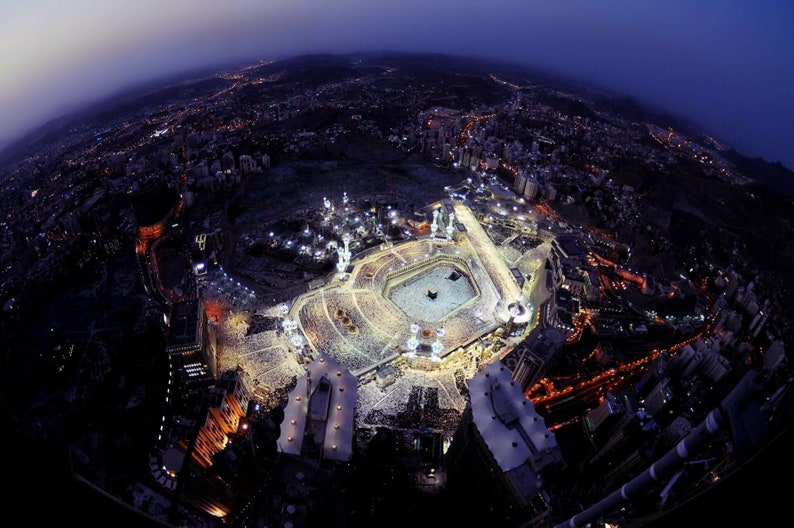 Op de afbeelding: Een nachtelijke luchtfoto van de Kaaba in Mekka, Saoedi-Arabi&euml;. De Kaaba is een kubusvormig gebouw dat de heiligste plaats in de islam is. Het is omgeven door een grote binnenplaats, en er zijn veel mensen verzameld eromheen. De stadslichten verlichten de sc&egrave;ne.