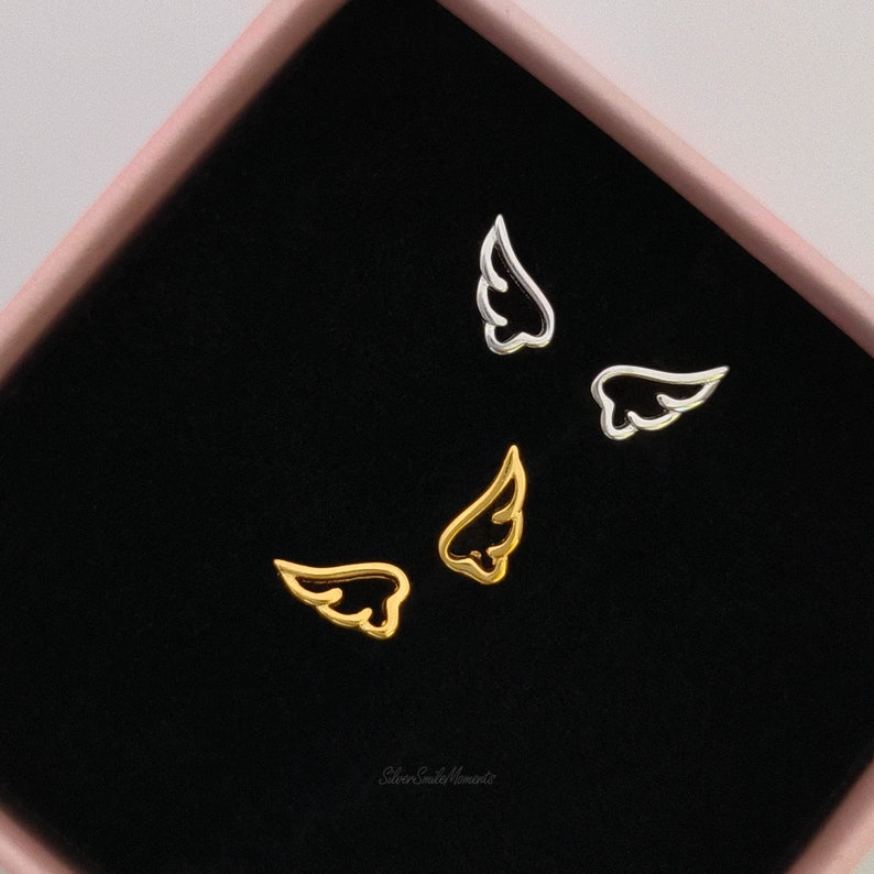 May include: A set of three silver and gold angel wing earrings in a black velvet box. The earrings are small and delicate, and they would make a great gift for a loved one.