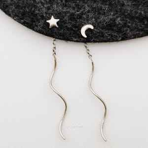 May include: A pair of silver dangle earrings with a star and crescent moon charm. The earrings have a wavy, curved design.