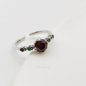 May include: Silver ring featuring a red gemstone surrounded by smaller green and blue gemstones. The ring is marked with "S925".