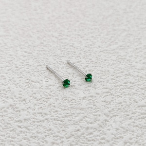 May include: A pair of small stud earrings with bright green gemstones. The earrings have a silver-colored post and setting. The gemstones are round and appear to be emeralds. The earrings are displayed on a textured, off-white surface.