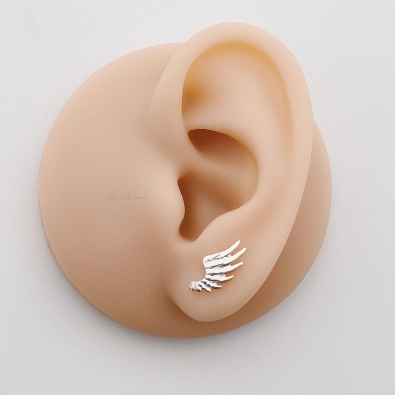 May include: A close-up of a silver wing earring on a model's ear. The earring is small and delicate, with a simple design.