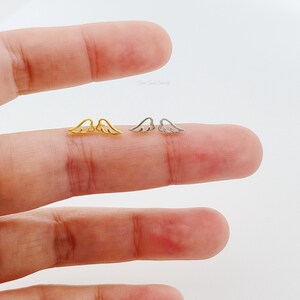 May include: A pair of small angel wing earrings, one pair is gold and the other is silver. The earrings are shown on a finger.