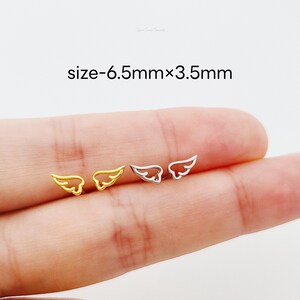 May include: A close-up of a pair of angel wing earrings. The earrings are 6.5mm x 3.5mm and are shown on a finger. One earring is gold and the other is silver.