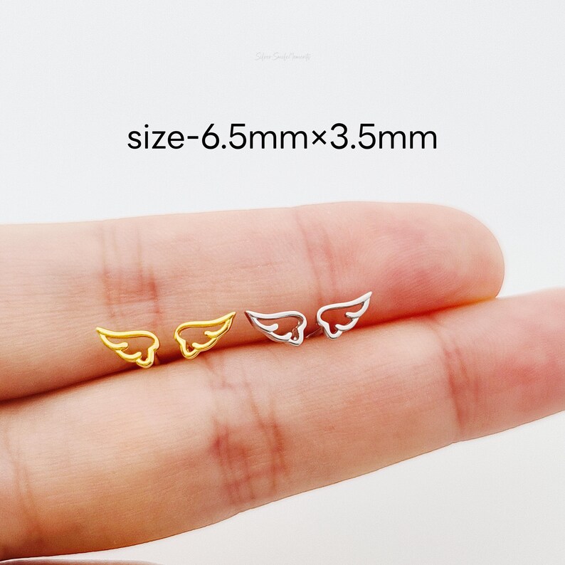 May include: A close-up of a pair of angel wing earrings. The earrings are 6.5mm x 3.5mm and are shown on a finger. One earring is gold and the other is silver.