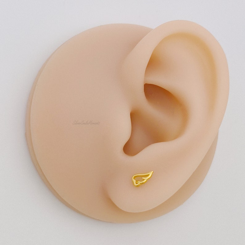 May include: Gold wing stud earring on a model ear. The wing is a simple design with a delicate outline.