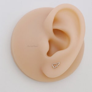 May include: A close-up of a fake ear with a small silver wing earring. The earring is a simple design with a delicate wing shape.
