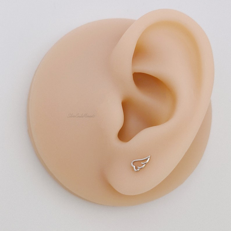 May include: A close-up of a fake ear with a small silver wing earring. The earring is a simple design with a delicate wing shape.