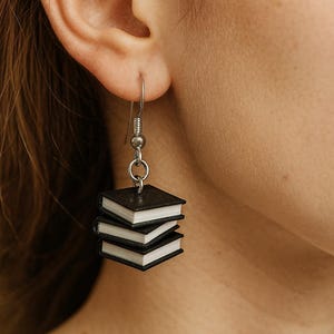 May include: A silver earring with a stack of three miniature black books with white pages. The earring is dangling from a person's ear, showcasing the unique, literary-themed jewelry.