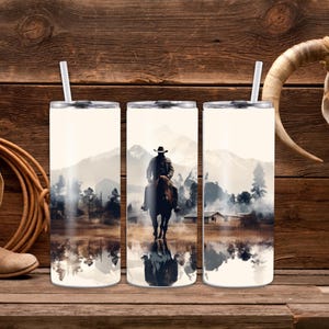 May include: Three stainless steel tumblers with a design of a cowboy riding a horse through a misty landscape. The design is reflected in the water below the horse.