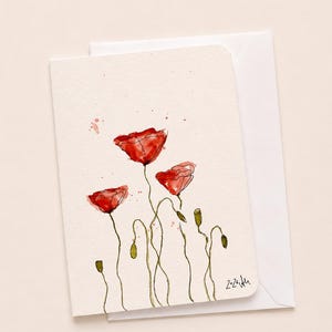 Könnte beinhalten: Eine Aquarell-Grußkarte mit roten Mohnblumen mit grünen Stielen und Knospen. Die Karte befindet sich auf einem cremefarbenen Hintergrund mit einem weißen Umschlag. Das Kunstwerk hat einen zarten, künstlerischen Stil.