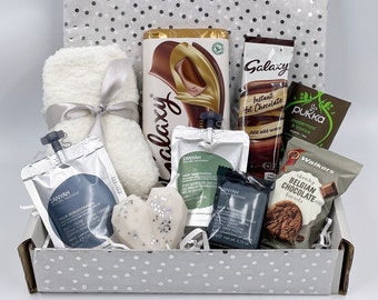 May include: A white gift box filled with various items, including a white towel, a heart-shaped candle, two tubes of body cream, a chocolate bar, a packet of hot chocolate mix, a packet of peppermint and licorice, and a packet of chocolate biscuits.