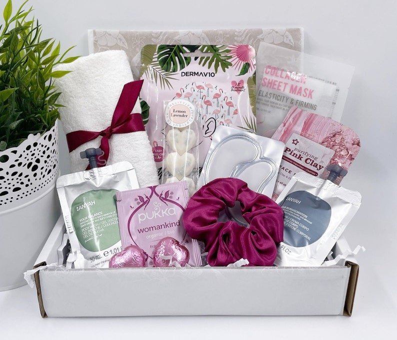 May include: A pink and white gift box filled with beauty products. The box contains a pink face mask, a pink clay mask, a white sheet mask, a white towel, a pink headband, a white bottle of face serum, a white bottle of body lotion, and a pink and white box of face masks.