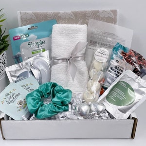 May include: A white gift box filled with various self-care items, including a white towel, face masks, eye masks, bath bombs, and a hair scrunchie. The box is filled with white shredded paper and has a white lid with a gray floral pattern.