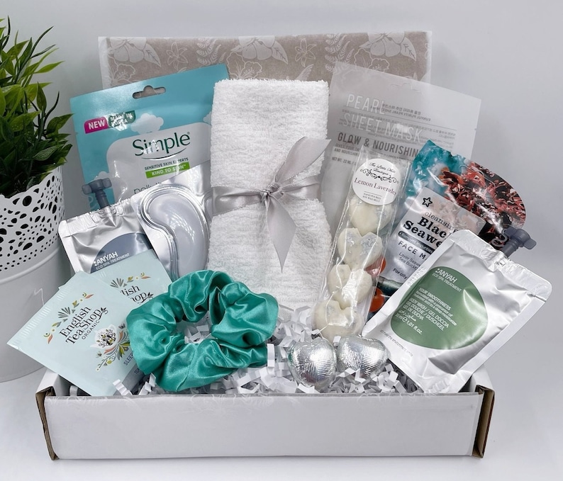May include: A white gift box filled with various self-care items, including a white towel, face masks, eye masks, bath bombs, and a hair scrunchie. The box is filled with white shredded paper and has a white lid with a gray floral pattern.