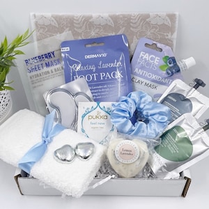 May include: A spa gift basket with a white towel, a blue hair scrunchie, a white soap bar with the text "Lemon Lavender", a silver heart-shaped chocolate, and various face and foot masks in clear plastic packaging.