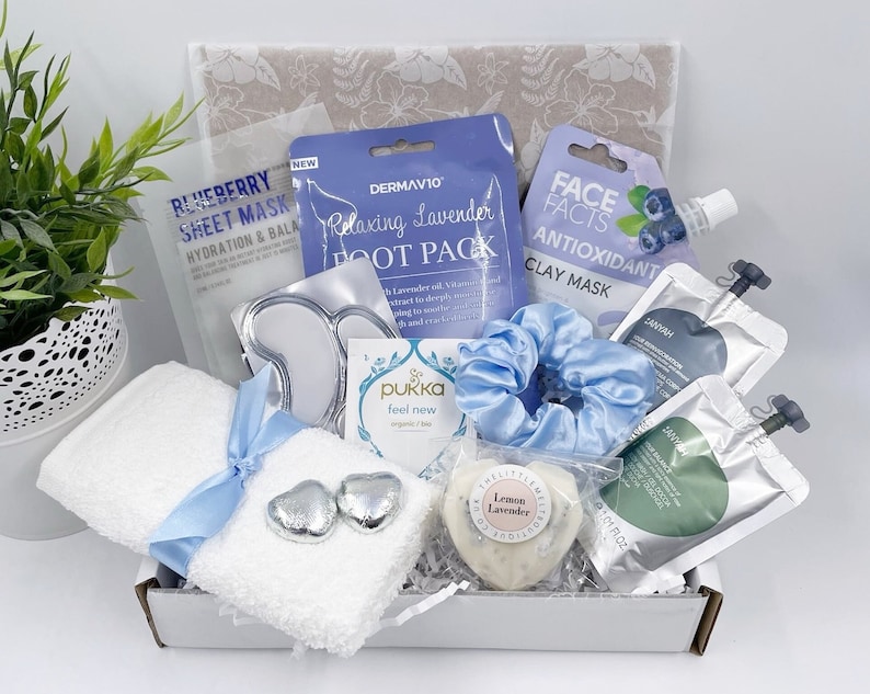 May include: A spa gift basket with a white towel, a blue hair scrunchie, a white soap bar with the text "Lemon Lavender", a silver heart-shaped chocolate, and various face and foot masks in clear plastic packaging.