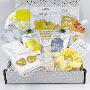 May include: A white gift box filled with self-care items, including face masks, a towel, bath bombs, and a hair scrunchie. The box is decorated with a silver and white polka dot pattern. The items are all in shades of yellow and white.