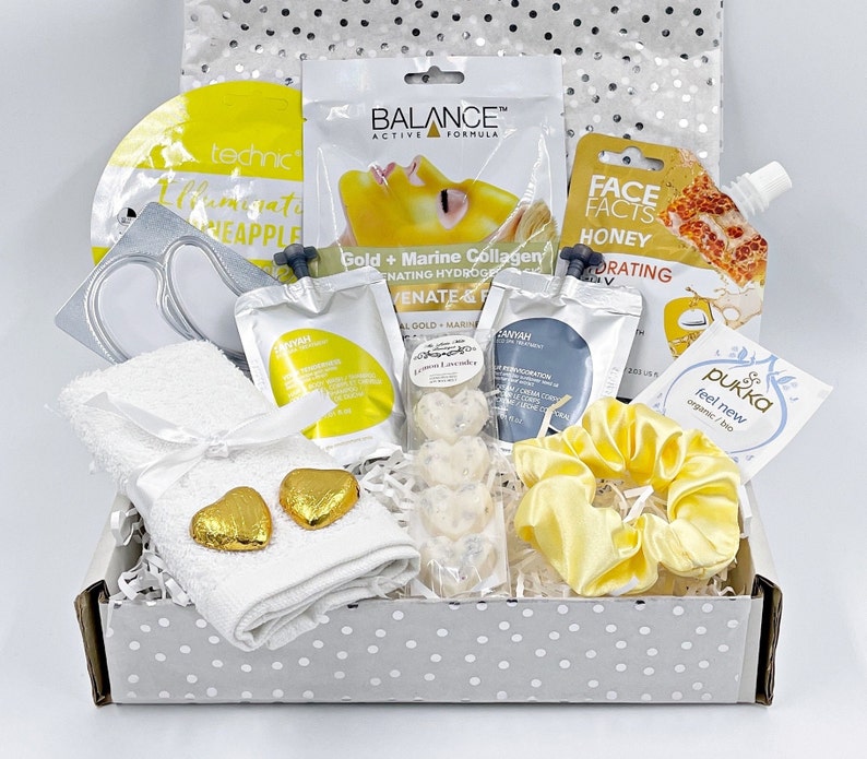 May include: A white gift box filled with self-care items, including face masks, a towel, bath bombs, and a hair scrunchie. The box is decorated with a silver and white polka dot pattern. The items are all in shades of yellow and white.