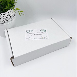 Pu&ograve; includere: Una scatola di spedizione rettangolare in cartone bianco con un adesivo rettangolare sulla superficie superiore. L'adesivo mostra illustrazioni di foglie verdi e il testo "Packed with Love Just for you" in un carattere scuro scritto a mano.