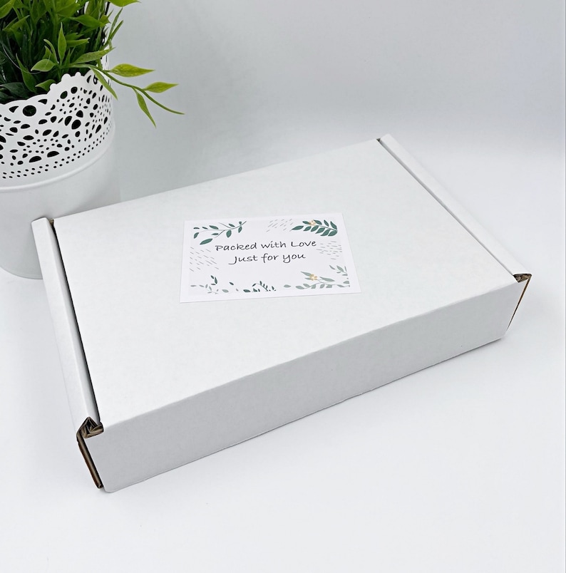 Pu&ograve; includere: Una scatola di spedizione rettangolare in cartone bianco con un adesivo rettangolare sulla superficie superiore. L'adesivo mostra illustrazioni di foglie verdi e il testo "Packed with Love Just for you" in un carattere scuro scritto a mano.