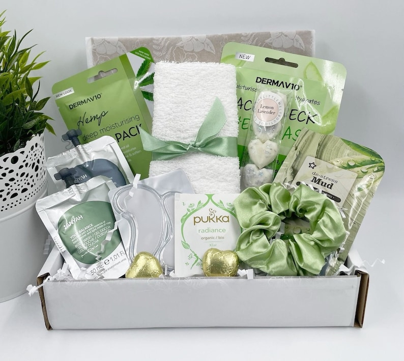 May include: A white gift box filled with green and white spa products. The box contains a white towel, face masks, a hair scrunchie, bath bombs, and chocolate hearts. The box is decorated with green and white tissue paper.