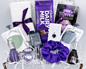 Può includere: Una scatola regalo piena di vari articoli, tra cui un asciugamano viola, una tavoletta di cioccolato Cadbury Dairy Milk, un elastico per capelli viola, una maschera per il viso, una collana con un ciondolo di pietra nera e una bottiglia di smalto per unghie.