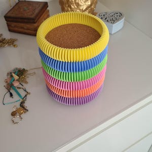 Può includere: Un set di sei sottobicchieri rotondi in cartone ondulato colorati. I sottobicchieri sono gialli, blu, verdi, rosa, arancioni e viola. Il sottobicchiere superiore ha una base in sughero.