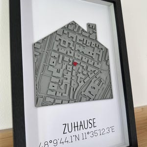 Puede incluir: Arte de mapa 3D enmarcado en un marco negro. El mapa gris, con forma de casa, tiene un marcador de corazón rojo. La palabra "ZUHAUSE" y las coordenadas están impresas debajo.