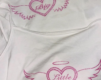 May include: Two white tank tops with pink glitter heart designs. One top says "Big" inside the heart with angel wings, and the other says "Little" with similar wings. The designs include a halo above the heart.