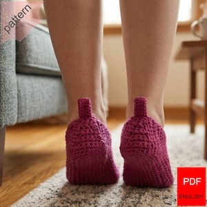 May include: A pair of magenta crochet slippers with a bottle-shaped design. The slippers have a textured appearance and a small loop at the back. The image is taken from a low angle, showing the slippers on a light-colored rug.
