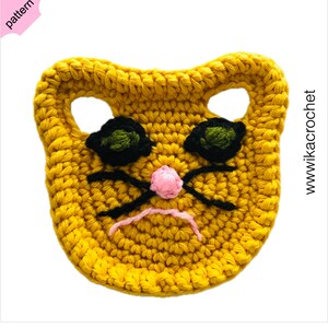Puede incluir: Una cara de gato amarilla tejida a crochet con ojos negros, una nariz rosa y bigotes negros. La cara del gato tiene dos agujeros para las orejas.