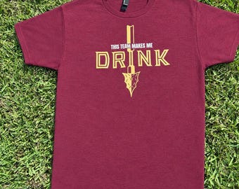 May include: A maroon short-sleeve t-shirt featuring a yellow graphic of an arrowhead pointing downward. The text "THIS TEAM MAKES ME" is printed in white above the word "DRINK" in large yellow letters, with the arrowhead integrated into the design.