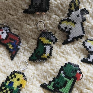 Puede incluir: Una colección de pendientes de pájaros en pixel art, que incluyen cacatúas, periquitos y otros loros coloridos. Cada pendiente está hecho de pequeñas cuentas de colores y presenta un gancho plateado. Los pendientes se muestran sobre una superficie texturizada de color crema.