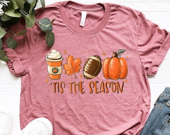 May include: A heather mauve t-shirt with a graphic design of a coffee cup, a leaf, a football, and a pumpkin. The text "Tis the Season" is printed below the graphic.