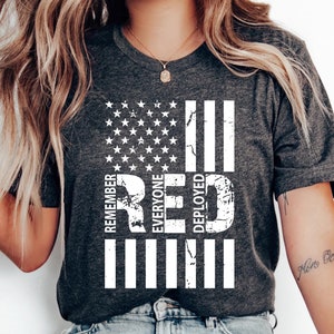 May include: A gray t-shirt with a distressed white American flag design. The flag has the words "RED" and "REMEMBER EVERYONE DEPLOYED" written vertically on it.