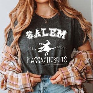 May include: A dark gray t-shirt with white text that reads "Salem Est. 1626. Massachusetts" and a white silhouette of a witch flying on a broomstick.