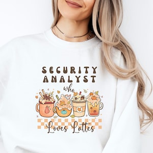 May include: White sweatshirt with the text "SECURITY ANALYST who loves Lattes" and a graphic of four fall-themed latte cups with autumn leaves and pumpkins.
