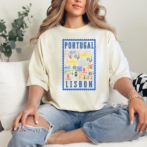 May include: Cream-colored t-shirt featuring a blue postage stamp design with the words "PORTUGAL LISBON". The stamp includes colorful illustrations of Portuguese landmarks and symbols. The shirt is worn by a person.