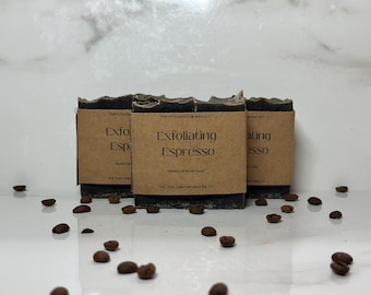 May include: Three dark gray exfoliating espresso soap bars with brown labels and coffee beans scattered around. The labels read "Exfoliating Espresso" in a simple font. The soap has a rough, textured top.