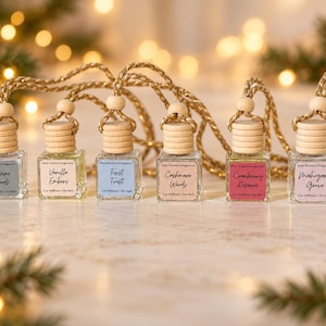 May include: Six small square glass car diffuser bottles with wooden caps and braided hanging cords. Each bottle features a label with the text Pure Essence Fragrance and scent names: Alpine Woods, Vanilla Embers, First Frost, Cashmere Woods, Cranberry Reserve, and Mahogany Grove.