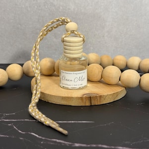Op de afbeelding: Een doorzichtig glazen autodiffuserflesje met een lichthouten dop en een beige en goudkleurig gevlochten ophangkoord. Het etiket vermeldt Pure Essence Fragrance LLC, Ocean Mist, Car Diffuser | Net 10 ml. Het flesje staat op een ronde houten schijf met houten kralen.