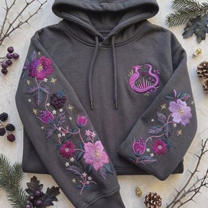 Puede incluir: Una sudadera con capucha gris carbón que presenta un intrincado bordado en ambas mangas y en el pecho. El bordado representa flores moradas y rosas, bayas oscuras y pequeñas estrellas doradas. El pecho muestra una serpiente y un ave.