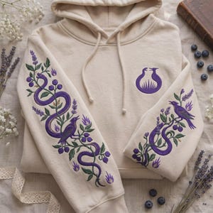 Könnte beinhalten: Ein beigefarbener Kapuzenpullover mit violetter Stickerei an beiden Ärmeln und auf der Brust. Die Ärmelmotive zeigen eine violette Schlange und einen Vogel, die mit grünen Ranken, Blättern und Beeren verziert sind. Ein violettes Emblem ist mittig.