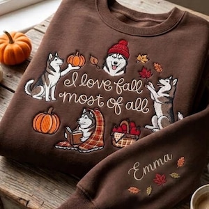 Pode incluir: Uma camisola castanha de gola redonda com bordados de três huskies, abóboras, um cesto de maçãs e folhas de outono. O texto diz I love fall most of all. A manga está bordada com o nome Emma e folhas coloridas.