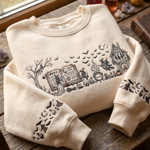 May include: A cream-colored crewneck sweatshirt featuring detailed black line art embroidery. The design depicts a witch reading a spellbook, a bubbling cauldron, a house, bats, and stars. The sleeves are decorated with matching witchy motifs.